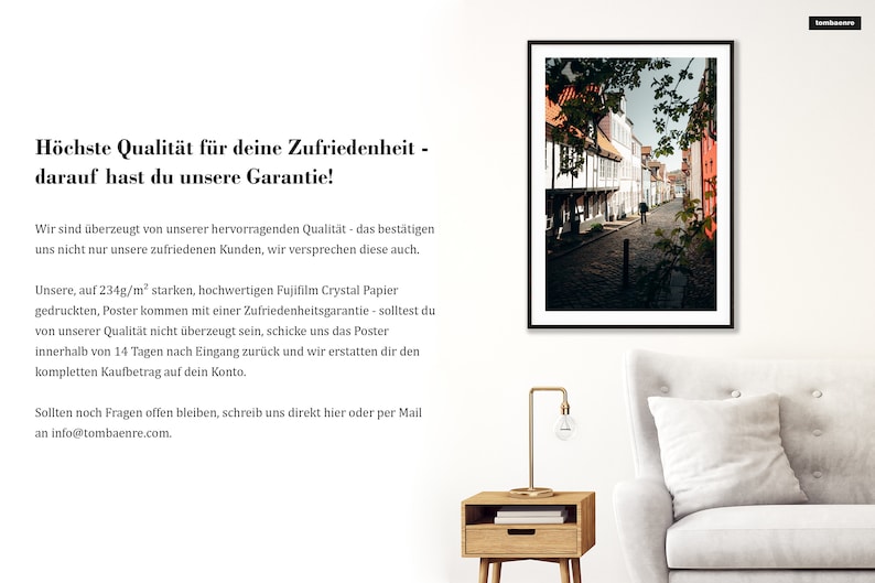 May include: A framed photograph of a narrow, cobblestone street lined with historic buildings. The buildings are a mix of white and red brick, with some featuring decorative timber framing. The street is empty except for a single person walking in the distance. The photograph is displayed on a white wall, with a wooden side table and a white sofa in the foreground.