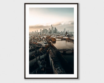 May include: An aerial view of a city skyline with a river running through it. The buildings are mostly tall and modern, with some older buildings in the foreground. The river is wide and calm, with a bridge crossing it in the distance. The sky is a soft blue with some clouds.