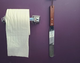May include: A roll of white toilet paper on a chrome holder, next to a novelty poop knife. The knife has a wooden handle and the word "POOP KNIFE" engraved on the blade. A wooden poop-shaped hook is above the knife.