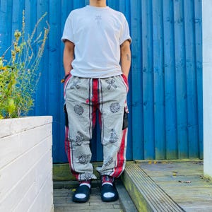 May include: A white short-sleeved t-shirt with a small graphic on the chest paired with grey cargo trousers featuring a repeating black floral pattern, vertical red and black stripes along the sides, a drawstring waist, and black slide sandals.