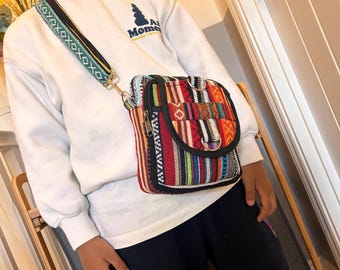 May include: A colorful crossbody bag with a patterned design. The bag features a variety of colors, including red, blue, green, and yellow. The strap is decorated with an embroidered design. The bag is rectangular with a flap closure and a zipper.