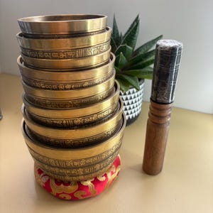 May include: A collection of golden singing bowls with etched inscriptions, stacked on a red and gold cushion. A wooden mallet with a wrapped handle is placed beside the bowls. A potted plant is in the background.