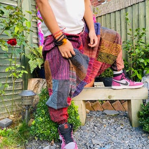 May include: Patchwork harem pants in shades of red, purple, blue, and brown. The pants have a drawstring waist and are worn with pink and black high-top sneakers. The pants are made of various fabric patches with different patterns.