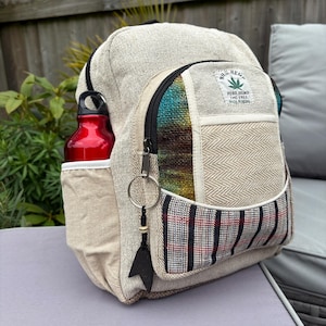 May include: A beige hemp backpack with a red water bottle in the side pocket. The backpack has a colourful front pocket with a black zip and a small keyring. The text "PURE HEMP THC FREE MADE IN NEPAL" is visible on the front.