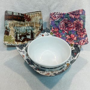 May include: A white ceramic bowl sits inside a fabric bowl cozy. Three fabric bowl cozies are shown: one with a brown and tan outdoor lodge theme, one with a purple floral pattern, and one with a white background featuring black food illustrations.
