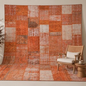 Puede incluir: Una alfombra rectangular grande con un diseño de patchwork en varios tonos de naranja, óxido y crema. La alfombra muestra un patrón desgastado y descolorido con segmentos rectangulares visibles, cubriendo tanto el suelo como la pared detrás de una silla.