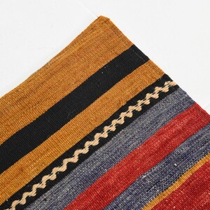 May include: A woven textile featuring a series of parallel stripes in mustard yellow, black, blue, and red. A thin, cream-colored zigzag line separates the black and blue sections, and a small portion of a geometric pattern is visible at the corner.