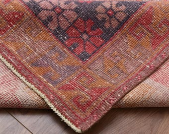 May include: Close-up of a rolled-up, vintage-style rug with a geometric pattern. The rug features a central design in red, purple, and orange hues, with a cream-colored border. The rug is made of woven material.