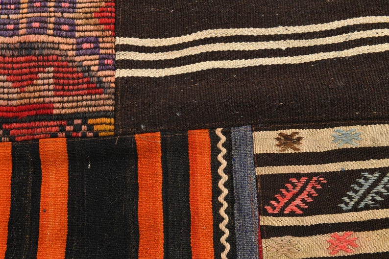 May include: A woven textile rug featuring a patchwork design of various patterns. Sections include black and white horizontal stripes, vibrant orange and black vertical stripes, and a multi-colored geometric grid with red, purple, and orange tones.
