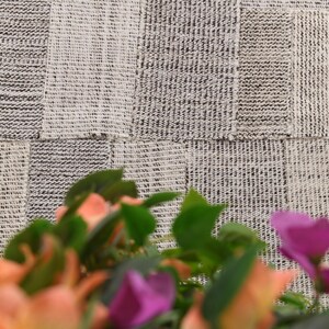 以下が含まれることがあります： グレーと白の色合いのパッチワークデザインの織物。明るい糸と暗い糸が混ざり合い、繊細な模様を作り出しています。前景には、オレンジと紫の色合いのぼやけた花があります。