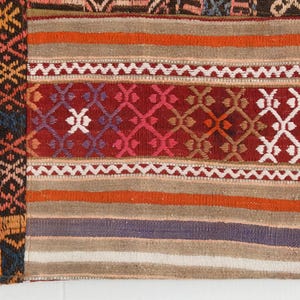 May include: A woven textile featuring a patchwork design with a dark section on the left displaying a blue, orange, and black diamond pattern. The right section consists of horizontal stripes in red, orange, beige, and purple with geometric motifs.