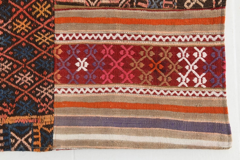 May include: A woven textile featuring a patchwork design with a dark section on the left displaying a blue, orange, and black diamond pattern. The right section consists of horizontal stripes in red, orange, beige, and purple with geometric motifs.