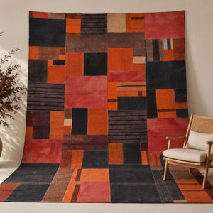 Op de afbeelding: Een groot vloerkleed met een geometrisch patchworkontwerp in tinten zwart, oranje, rood en crème. Het patroon bestaat uit diverse rechthoekige en vierkante blokken, sommige met effen kleuren en andere met horizontale lijnen.
