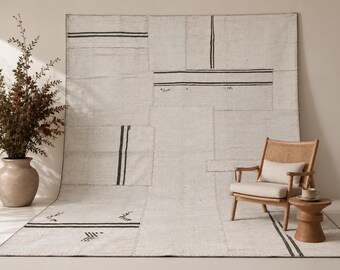 May include: A large, off-white area rug featuring a patchwork design with black linear stripes and small geometric motifs. The rug is displayed vertically against a wall, accompanied by a wooden chair with a cane back and a small wooden side table.