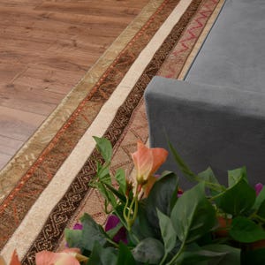 Pode incluir: Uma secção de um tapete estampado com tons de castanho, bege e vermelho, ao lado de um sofá cinzento. O tapete tem um design listrado com padrões geométricos. Um buquê de flores com folhas verdes e flores laranja está em primeiro plano.