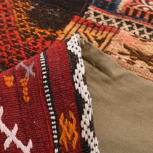 May include: A woven textile pillow cover featuring a folded corner that reveals a solid olive green fabric backing. The front side displays a complex geometric pattern in shades of deep red, black, orange, and white with intricate stitched details.