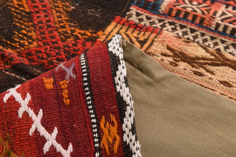 May include: A woven textile pillow cover featuring a folded corner that reveals a solid olive green fabric backing. The front side displays a complex geometric pattern in shades of deep red, black, orange, and white with intricate stitched details.