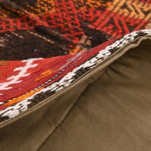 May include: A decorative pillow cover featuring a woven textile front with geometric patterns in shades of red, orange, brown, and white. The back is made of solid olive green fabric, and a zipper closure is visible along the edge of the cover.