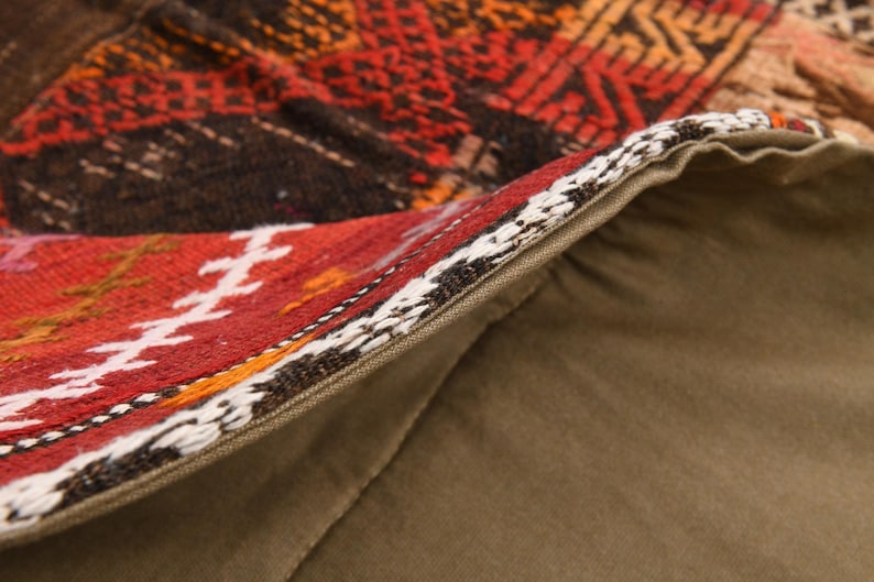 May include: A decorative pillow cover featuring a woven textile front with geometric patterns in shades of red, orange, brown, and white. The back is made of solid olive green fabric, and a zipper closure is visible along the edge of the cover.