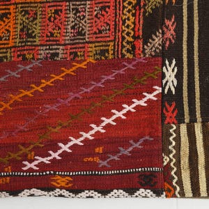 May include: A woven textile rug featuring a patchwork design with various geometric patterns. The color palette includes deep red, dark brown, orange, and cream. Visible details include diagonal cross-stitching and striped borders.