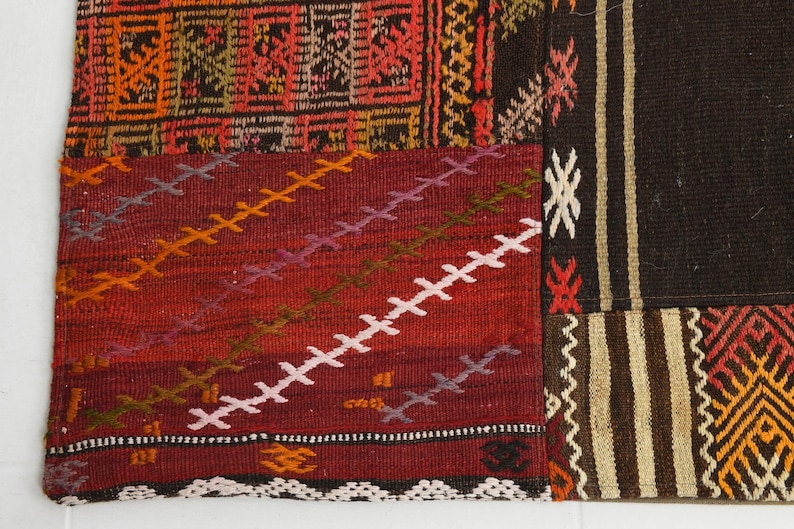 May include: A woven textile rug featuring a patchwork design with various geometric patterns. The color palette includes deep red, dark brown, orange, and cream. Visible details include diagonal cross-stitching and striped borders.