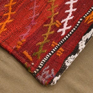 May include: A woven textile corner featuring a deep red base color with vertical stripes of embroidered cross-stitch patterns in orange, purple, olive green, and white. The fabric has a textured, coarse weave with a distinct geometric border.