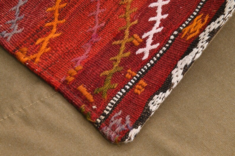 May include: A woven textile corner featuring a deep red base color with vertical stripes of embroidered cross-stitch patterns in orange, purple, olive green, and white. The fabric has a textured, coarse weave with a distinct geometric border.