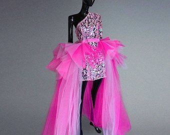 May include: A one-shoulder, sequined mini-dress in pink and silver, with a layered tulle skirt and a long, flowing train. The dress is displayed on a black mannequin against a gray background. The dress features a bow at the waist.