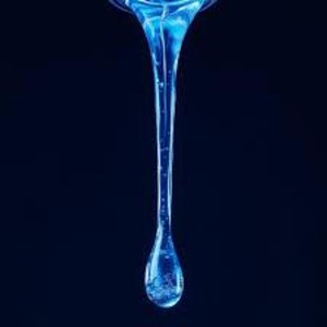 May include: A close-up of a single, elongated, blue liquid drop against a dark blue background. The drop is suspended mid-air, with a narrow stream flowing into a larger, tear-shaped droplet at the bottom. The liquid appears translucent, with subtle internal textures.