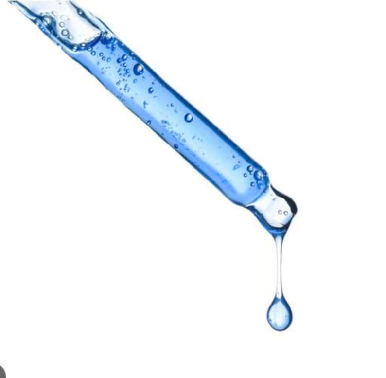 May include: A close-up of a blue glass dropper with a clear liquid inside and a single drop of liquid suspended from the tip. The dropper is angled diagonally against a white background.