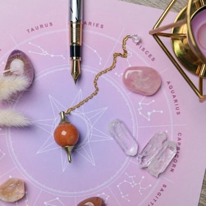 Könnte beinhalten: Ein Stillleben mit Stift, Pendel und Kristallen auf einer lila Oberfläche. Das Pendel hat eine orangefarbene Kugel und goldfarbene Akzente. Kristalle sind Bergkristall und Rosenquarz.