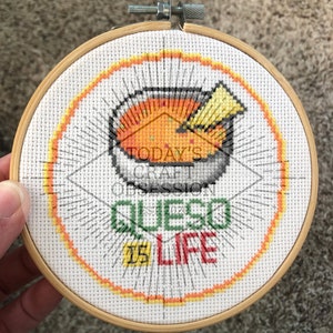 Puede incluir: Un aro de bordado blanco con un diseño de punto de cruz colorido que presenta un tazón de queso con una tortilla encima. El texto "Today's Craft Obsession Est. 2020 Queso Is Life" está bordado en hilo de colores.