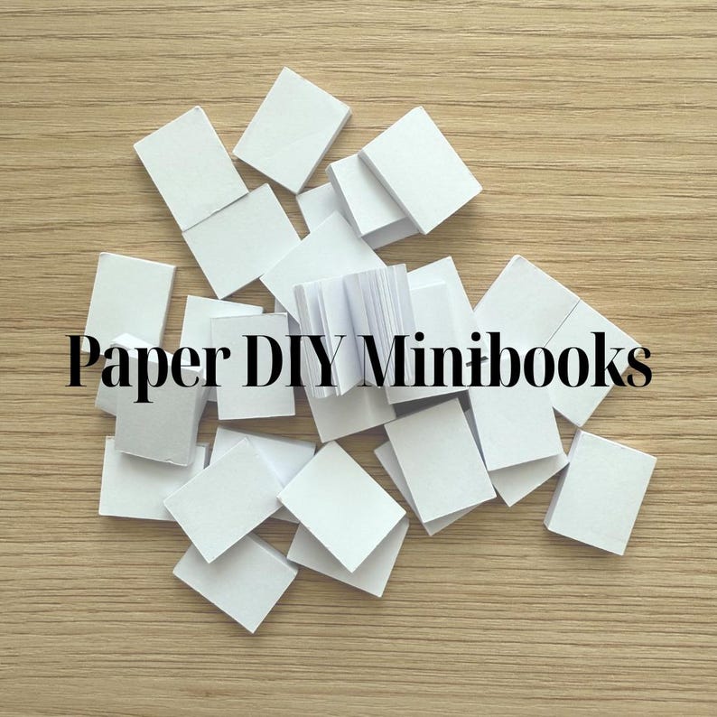 May include: A scattering of small, white paper rectangles and a tiny book on a wooden surface. The text "Paper DIY Minibooks" is displayed in black font, indicating a craft project or DIY kit for making miniature books.