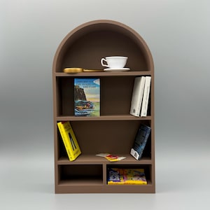 Puede incluir: Una estantería marrón en forma de arco con varios estantes. Contiene una taza de té y un platillo blancos, una lupa, varios libros y una pequeña tarjeta amarilla. Los libros incluyen títulos como "The House on the Cliff" y "Roald Dahl".