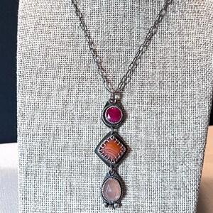 May include: A silver-toned necklace featuring a vertical pendant with three distinct stones. The top stone is a round, faceted deep pink gem, the middle is a square, banded orange agate, and the bottom is a pear-shaped, faceted pale pink stone.