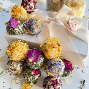 May include: A selection of floral bath bombs, decorated with dried petals in various colours. The bombs are presented on a white box tied with a cream ribbon. A small glass jar with a cork and a natural sponge are also visible.