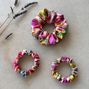 Puede incluir: Tres scrunchies de diferentes tamaños con estampado floral de colores. Los scrunchies están hechos de tela y tienen un aspecto fruncido.