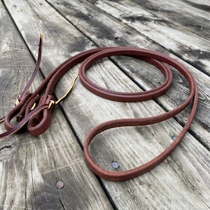 May include: A brown leather rein with a knotted end. The rein is coiled on a wooden surface.