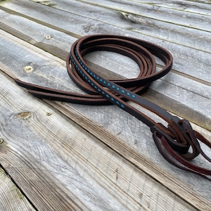 May include: A pair of brown leather reins with blue stitching. The reins are coiled together and have a decorative knot at the end.