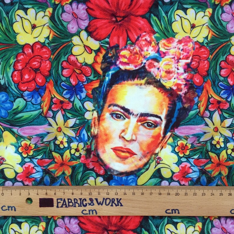 Op de afbeelding: Een kleurrijke stof met een bloemenpatroon en een portret van Frida Kahlo in het midden. De stof is groen, rood, geel, blauw en roze. Het portret is een close-up van Frida Kahlo's gezicht, met haar kenmerkende monobrauw en bloemenkrans.
