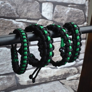 May include: Four black and green paracord bracelets with adjustable closures. The bracelets are displayed on a wooden stand.