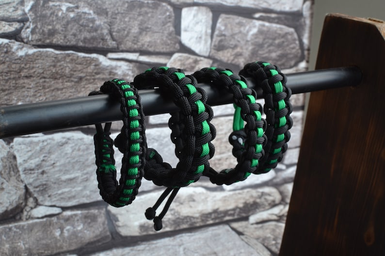 May include: Four black and green paracord bracelets with adjustable closures. The bracelets are displayed on a wooden stand.