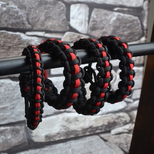 May include: Four black and red paracord bracelets, woven with a cobra stitch, are displayed on a black metal rod.