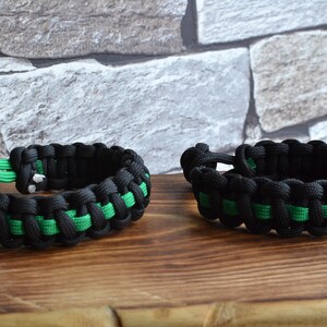 May include: Two black and green paracord bracelets with a woven design. The bracelets are on a wooden surface.