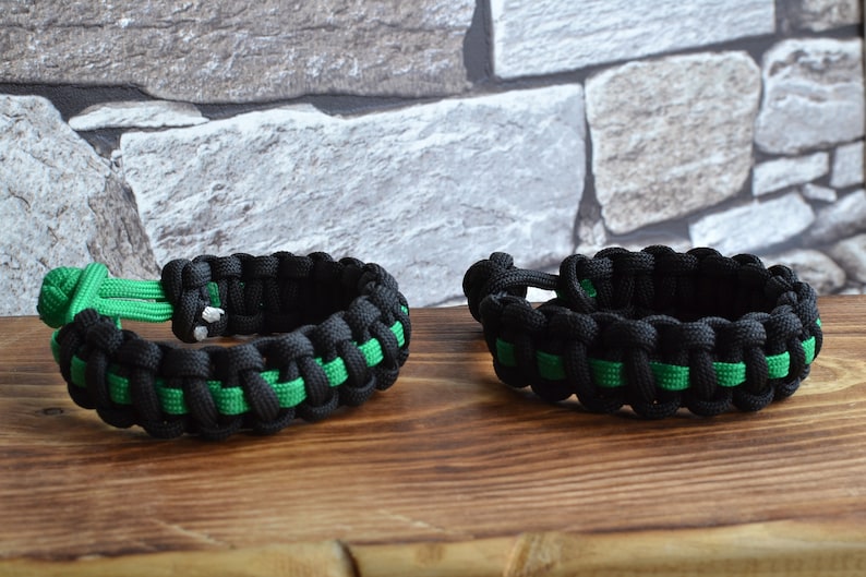 May include: Two black and green paracord bracelets with a woven design. The bracelets are on a wooden surface.