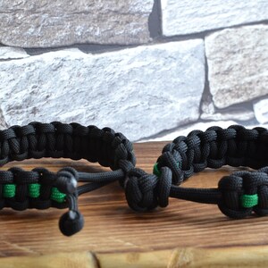 May include: Two black paracord bracelets with green accents. The bracelets are tied together with a knot.
