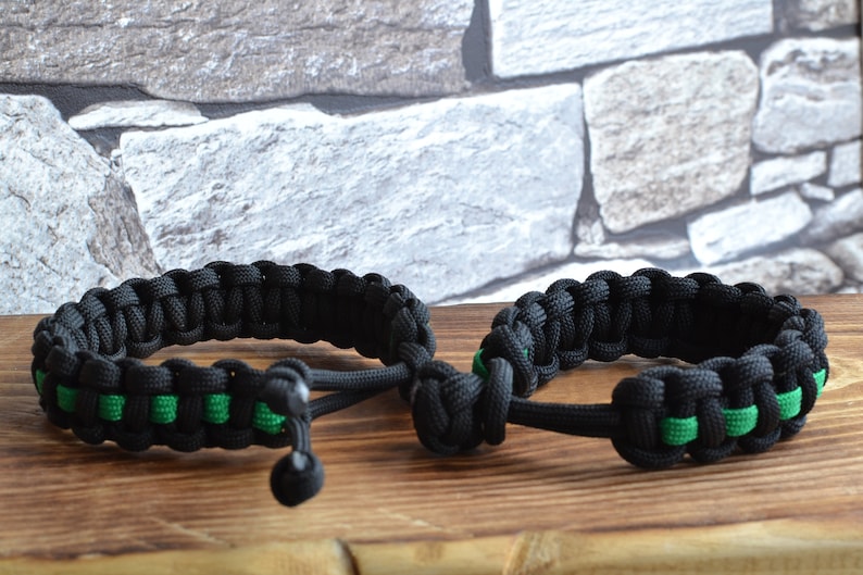 May include: Two black paracord bracelets with green accents. The bracelets are tied together with a knot.