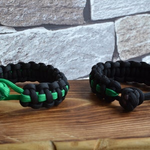 May include: Two black and green paracord bracelets with a knot on each end. The bracelets are on a wooden surface.