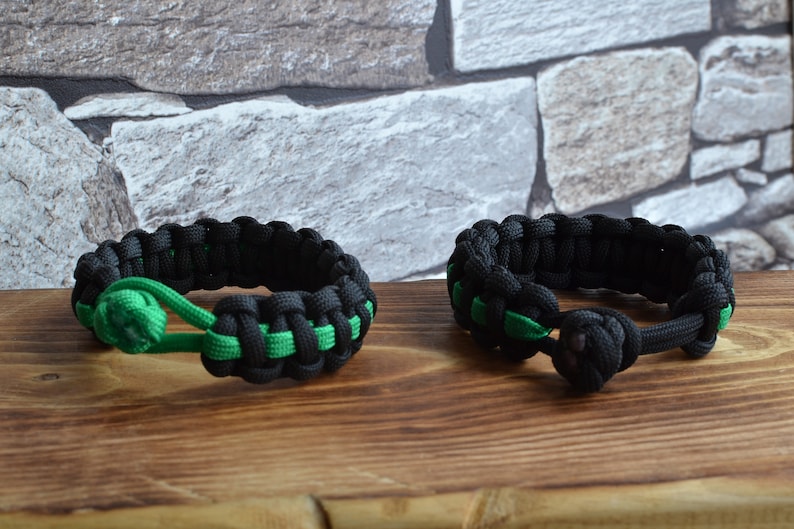 May include: Two black and green paracord bracelets with a knot on each end. The bracelets are on a wooden surface.