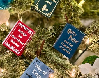 May include: Miniature book ornaments suspended on a Christmas tree. The ornaments feature titles such as "A Christmas Carol," "Rudolph the Red-Nosed Reindeer," "Harry Potter and the Chamber of Secrets," and "Way of Kings."